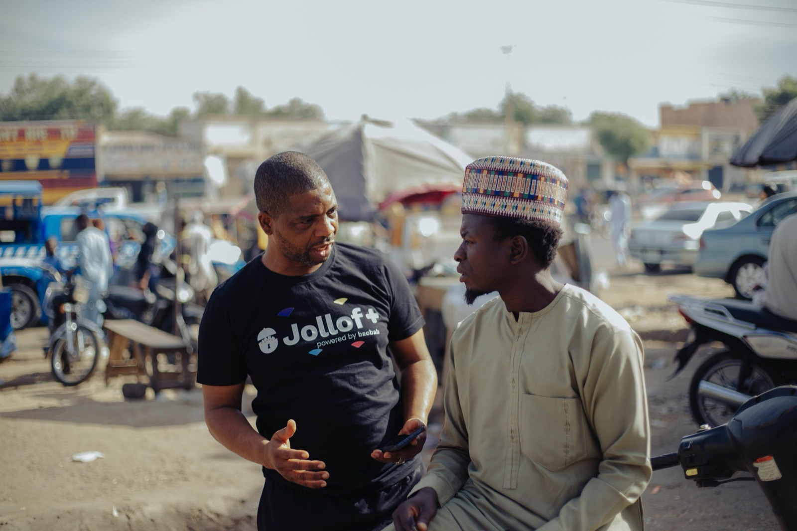 Two men in conversation at a market, one holding a phone
