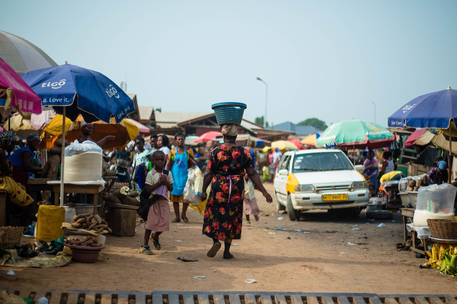 A bustling Ghanaian market street with umbrellas and a taxi